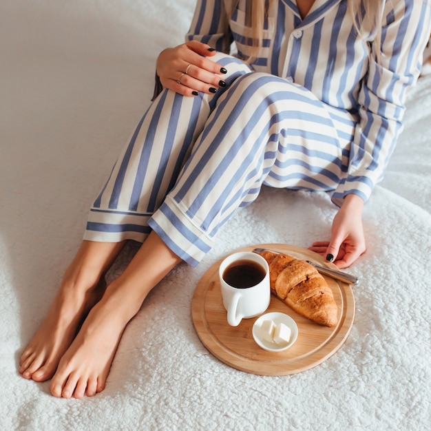 Frauenbeine im Pyjama und neben einem Kaffee mit einem Croissant