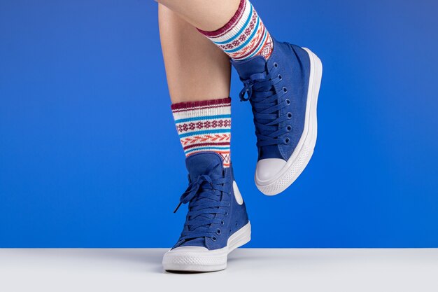 Frauenbeine in den blauen Turnschuhen und in den farbigen Socken mit Verzierungen, blauer Hintergrund. Sport und Erholung.