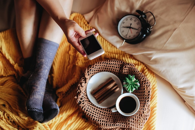 Frauenfüße auf Socken, die auf dem Bett mit Smartphone und Kaffee zum Frühstück entspannen