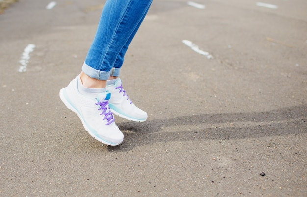 Frauenfüße in Blue Jeans und weißen Turnschuhen auf dem Asphalt