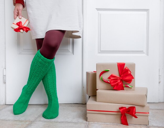 Frauenfüße in grünen Socken stehen in der Nähe der weißen Tür mit Geschenken am Vorabend der Feiertage Weihnachten und Neujahr.