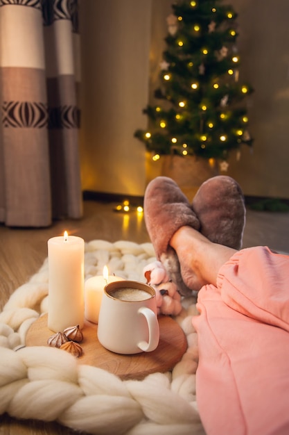 Frauenfüße in Hausschuhen aus Pelz, Tasse Cappuccino, Kerzen auf einer Decke aus dickem Garn und Weihnachtsbaum mit Lichtern. Die Atmosphäre von Wohnlichkeit und Behaglichkeit