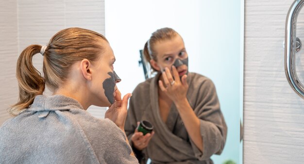 Frauengesundheit. Spa und Wellness. Frau in grauen Bademänteln mit Gesichtsmaske im Badezimmer mit Blick auf den Spiegel