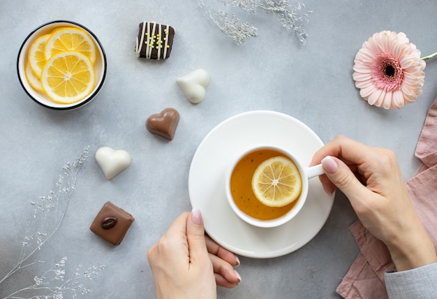 Frauenhände, die eine Tasse heißen Tee mit Zitrone und verschiedenen Süßigkeiten auf einer grauen Oberfläche mit Blumen halten.