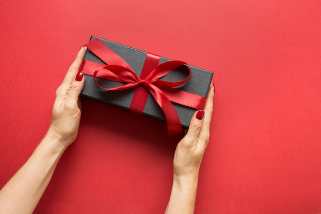 Frauenhände, die schwarze Geschenkbox halten, gewickelt mit rotem Band auf roter Oberfläche. Valentinstagskarte.