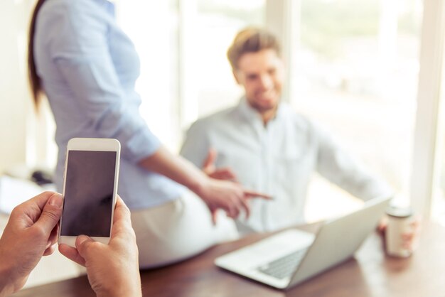 Frauenhände halten ein Smartphone im Hintergrund. Junges Geschäftspaar verwendet Laptop, spricht und lächelt, während es im Büro arbeitet