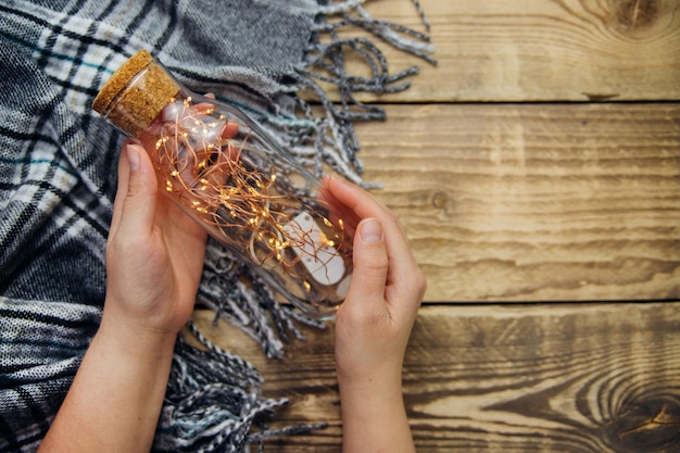 Frauenhände halten eine Glasflasche mit einer Girlande aus Weihnachtslichtern. Warmer grauer Schal oder Plaid auf einem hölzernen Hintergrund. Eine Postkarte mit Weihnachtsstimmung. Draufsicht mit Platz für Text.