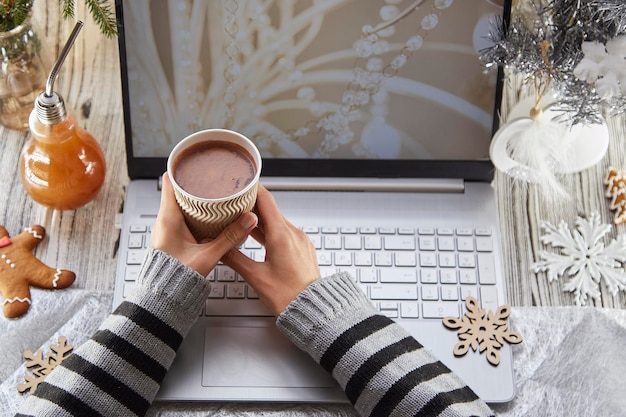 Frauenhände halten eine Papptasse Kakao Festliches Home Office Laptop Lebkuchen und Weihnachtsbaum mit Dekorationen Konzept des Webinars für Remote-Arbeit oder das Anrufen von Familienfreunden wegen Sperrung