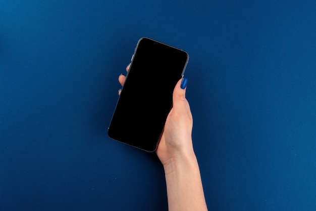 Foto frauenhände halten smartphone mit schwarzem bildschirm gegen klassischen blauen hintergrund, draufsicht