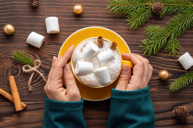 Frauenhände im Pullover halten eine Tasse Latte mit Marshmallows auf Holztisch