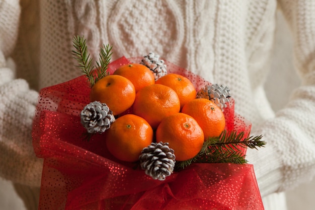 Frauenhände im weißen Pullover mit Blumenstrauß aus Mandarinen und Weihnachtsbaumasten. Essbares Bouquet von Früchten des neuen Jahres. Geschenk zu Weihnachten. DIY-Geschenk. nützliches Geschenk aus Früchten. Dekor von Früchten.