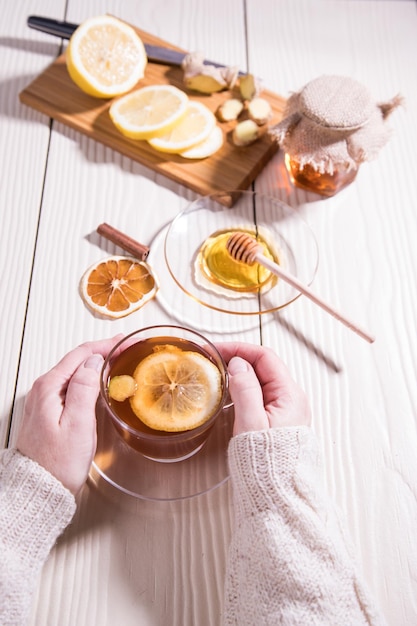 Frauenhände in einem weißen Strickpullover halten eine Tasse Vitamintee mit Zitronenhonig und Ingwer