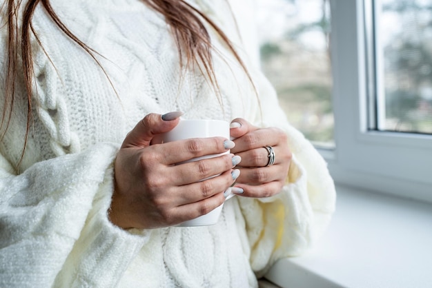 Frauenhände in warmweißem Winterpullover sitzen zu Hause auf der Fensterbank