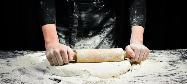Frauenhände mit einem Nudelholz bereiten den Teig für Brot oder Pizza zu Schwarzer Kochhintergrund