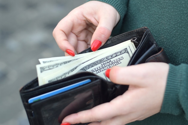 Frauenhände mit roten Nägeln halten schwarze Herrengeldbörse mit vielen US-Dollar-Scheinen. Konzept des Gehaltsverdienstes oder des Geldzählens