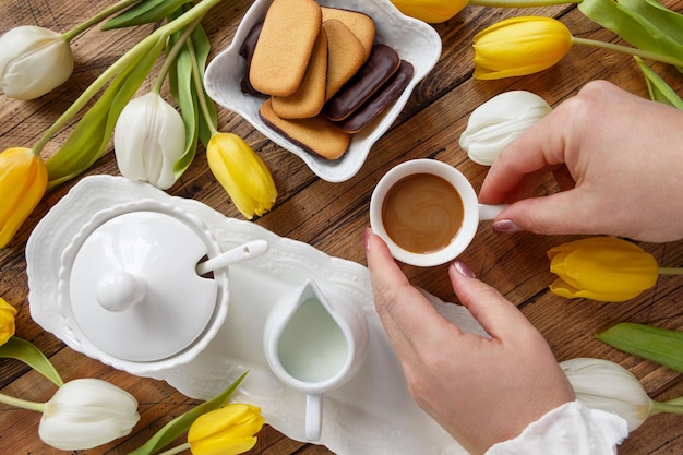 Frauenhände nehmen eine Kaffeetasse von einem Tablett zwischen weißen und gelben Tulpen auf Holztisch-Draufsicht. Romantisches Frühlingsfrühstück und Valentinstagskonzept