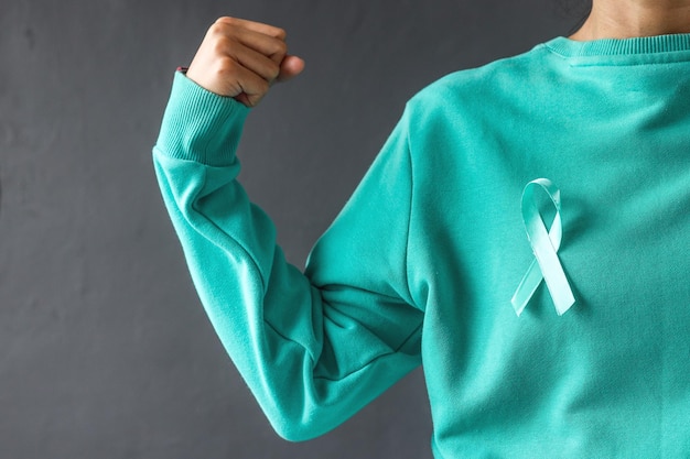 Frauenhände tragen ein blaugrünes T-Shirt und ein blaugrünes Bewusstseinsband und gestikulieren mit der starken Hand. Unterstützen Sie Krebs