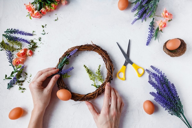 Frauenhänden machen Ostern Kranz aus Blumen, Kräutern und Eiern auf grau