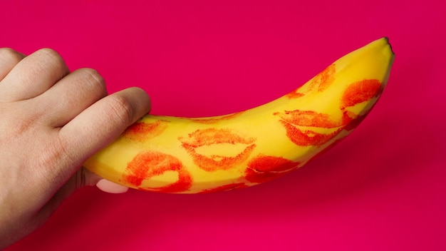 Frauenhand, die Banane mit roten Lippenstiftmarkierungen auf einem rosa Hintergrund hält