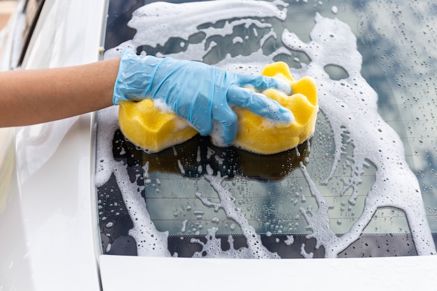 Frauenhand, die blaue Handschuhe mit dem gelben Schwamm wäscht modernes Auto des hinteren Spiegels trägt