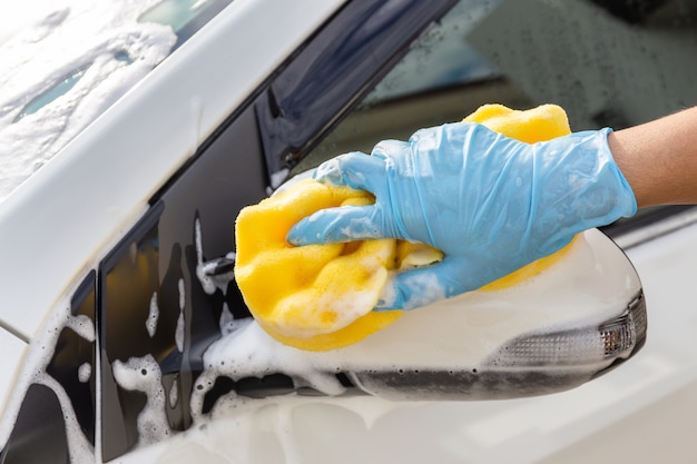 Frauenhand, die blaue Handschuhe mit waschendem modernem Auto des Seitenspiegels des gelben Schwammes trägt