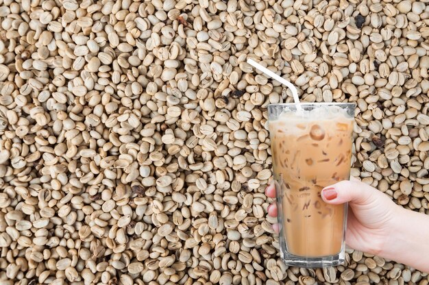 Frauenhand, die das Glas Eiskaffee auf ungerösteten Kaffeebohnen Hintergrund hältIced Latte Coffee
