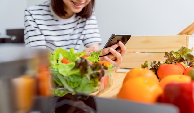 Frauenhand, die ein Smartphone und eine Salatschüssel mit Tomate und verschiedenem grünem Blattgemüse auf dem Tisch zu Hause hält, nehmen Sie Ihre Werbung.