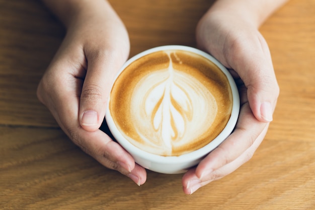Frauenhand, die eine weiße Kaffeetasse hält. Kaffee ist ein Latte. Tisch auf dem Holz