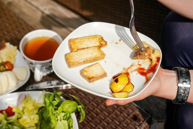 Frauenhand, die Gabel und Teller mit Grillkartoffeln und Tofu-Sojabohnenquark mit Ketchup und Senf hält - vegetarisches Grillen.