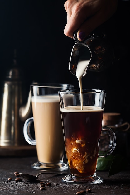 Frauenhand, die Milch in Glasschale des Kaffees auf schwarzer Oberfläche gießt