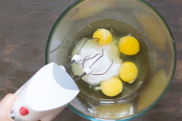 Frauenhand, die Mixer in der Glasschüssel mit Zucker und Eiern hält, bevor sie schlagen