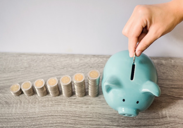 Frauenhand, die Münze in blaues Sparschwein mit Münzstapel-Wachstumsdiagramm steckt, Geld für zukünftige Investitionspläne und Pensionsfondskonzept sparend