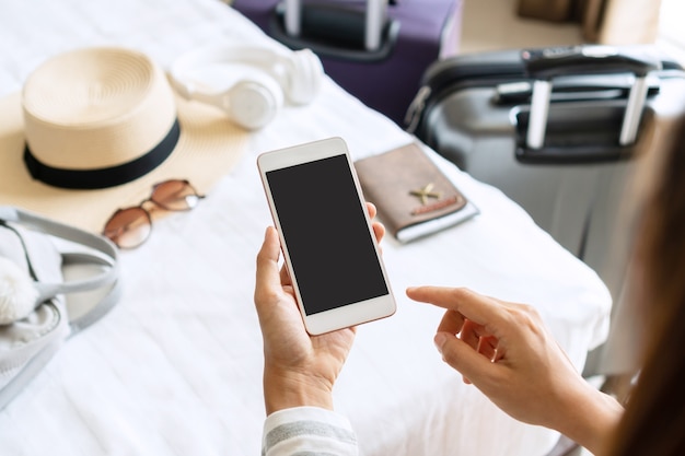 Frauenhand, die Smartphone mit Reiseaccessoires auf Bett im Hotelzimmer hält. Reise-, Entspannungs-, Reise-, Reise- und Urlaubskonzepte. Draufsicht und Kopierraum.