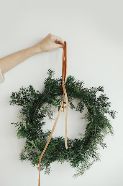 Frauenhand, die stilvollen Weihnachtskranz mit Samtband und Glocken auf weißer Wand hältUrlaubsdekor