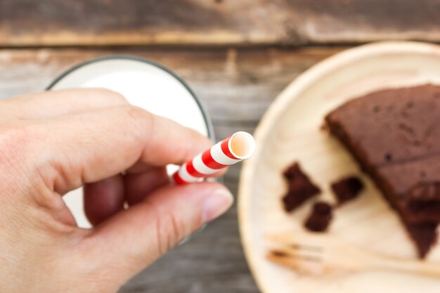 Frauenhand, die Stroh im Glas Milch hält. Schokoladenkuchen auf hölzerner Platte