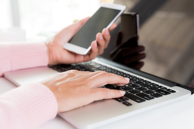 Frauenhand, die weißen Handy auf einer Tabelle mit einem Laptop im Büro hält