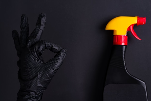 Frauenhand in einem schwarzen Latexhandschuh zum Reinigen, der ein OK-Zeichen und eine Plastiksprühflasche auf dem gleichen Farbhintergrund macht.