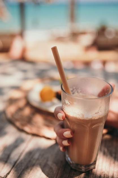 Frauenhand mit gelber Maniküre hält Eiskaffee Latte in hohem Glas mit Strohhalm am Meer in Strand b