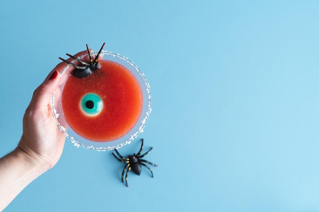 Frauenhand mit rotem Cocktailglas mit Halloween-Dekoration auf blauem Hintergrund Kopierbereich Vertikales Foto