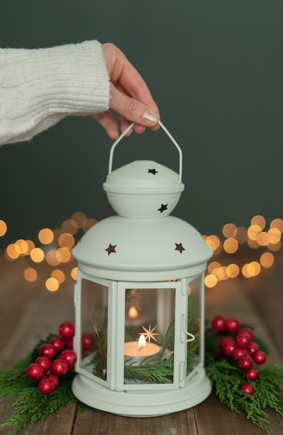 Frauenhand mit weißem Ärmel, der weiße Laterne mit Ornamenten und Weihnachtsbeleuchtung auf rustikalem Holztisch hält. Platz kopieren.