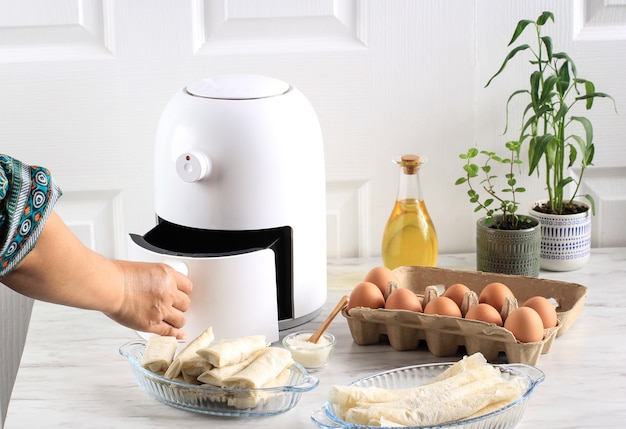 Frauenhand öffnen Airfryer-Behälter. Eine weiße Fritteuse oder eine ölfreie Fritteuse, eine Zange, eine klare Backform und ein Eiertablett stehen auf dem Holztisch in der Küche mit einer kleinen Pflanze im Topf (Luftfritteuse)