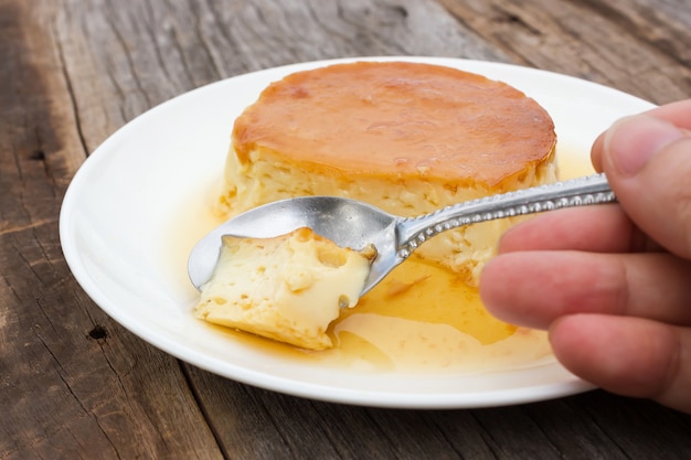 Frauenhandgebrauchslöffel, der selbst gemachten Karamellvanillepuddingpudding auf weißer Platte schneidet.