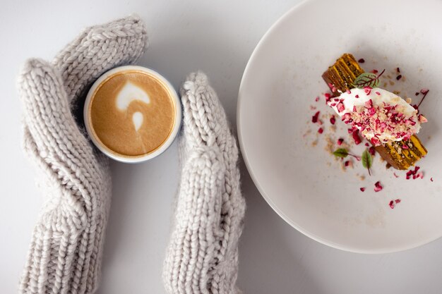 Frauenhandschuhe halten eine Kaffeetasse und einen Kuchen daneben auf einem weißen Tisch in der Nähe. Draufsicht. Weihnachtshintergrund. Konzept von Winter, Wärme, Feiertagen, Ereignissen.