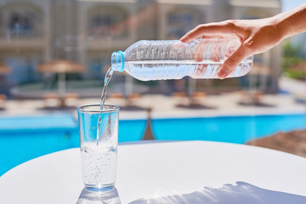 Frauenhandströmendes Wasser in einem Glas von der Plastikflasche agains Swimmingpoolhintergrund