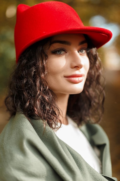 Frauenherbstporträt eines schönen, stilvollen Mädchens mit lockigem Haar in einem modischen Pullover mit grünem Schal und einem modischen roten Hut in einem Herbstpark im Sonnenlicht