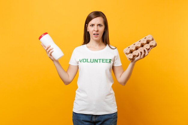 Frauenporträt im weißen T-Shirt mit schriftlicher Inschrift grüner Titel Freiwilliger halten Plastikflasche, Karton isoliert auf gelbem Hintergrund. Freiwillige kostenlose Hilfe, Müllsortierkonzept.