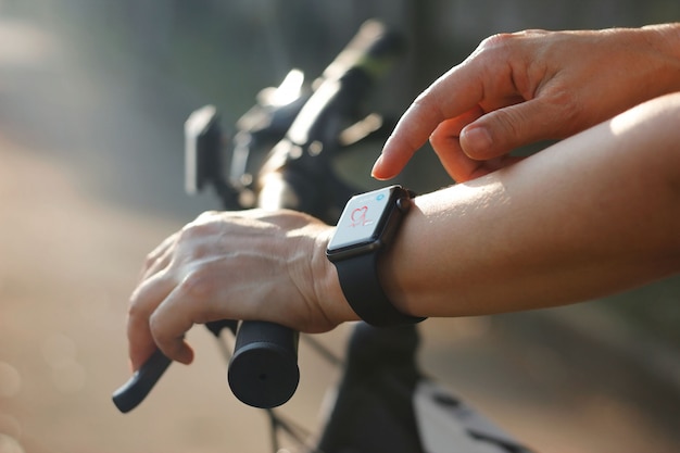 Frauenradfahrerhände mit Herzfrequenzikone auf intelligenter Uhr, Fahrrad an der Sommerspur