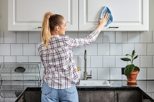 Frauenreinigungsküche mit Sprühreiniger und Spültuch, Konzept für Hygiene. blonde Frau in Freizeitkleidung, die moderne Küche säubert. Hausarbeit. Reinigung nach Hause. Rückansicht