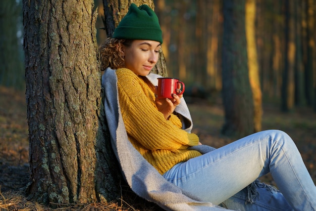 Frauenreisende genießen den Sonnenuntergang nach der Wanderung im Herbstwald, sitzen am Baum mit einer Tasse heißem Tee oder Kaffee