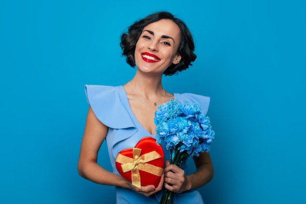 Frauentag St. Valentinstag Nahaufnahme Porträt einer eleganten, modisch lächelnden Frau im Kleid mit Blumenstrauß und Geschenkbox in den Händen isoliert auf blauem Hintergrund